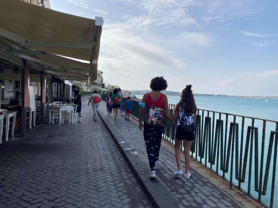 Il gruppo prosegue la passeggiata sul marciapiede che costeggia il mare. Un ringhiera in ferro sulla destra protegge il cammino, a sinistra i tavolini dei ristoranti all'aperto